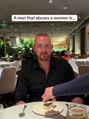 A man in a black shirt is seated at a restaurant table. The shot is centered, with the man looking at the camera. A hand enters the frame from the right, apparently preparing a dish on the table. The background shows other tables and decor of the upscale restaurant, with warm lighting.