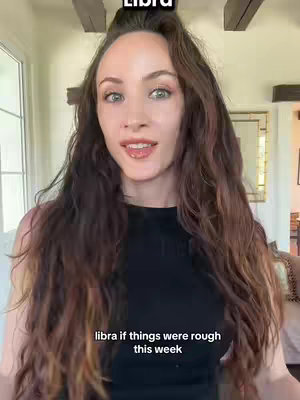 A woman with long, wavy brown hair is centered in a medium shot, facing the camera. She wears a black top. Text overlays appear at the top stating "LIBRA" and at the bottom, in a smaller and more informal font, "libra if things were rough this week". The background is a blurred interior setting, with light streaming in from a window. Overall, the image has a natural and inviting ambiance.