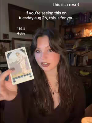 A woman with long dark hair holds up a tarot card in a room with books and lighting visible in the background. Text overlays describe a message.