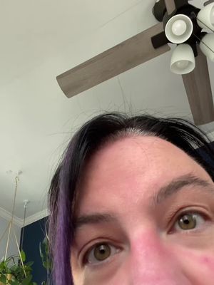 An eye level shot of a woman's face, angled slightly upwards. The focus is on her eyes, which are a light green color. The top of her forehead and a portion of her face, showing reddish skin. Her dark hair is parted and has a purple streak.