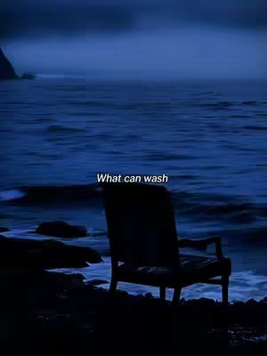 A nighttime scene at the beach with a dark silhouette of a chair on the shoreline. The sky is dark blue. The water is slightly visible.