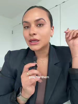 A woman with dark hair and medium skin tone is centered in the frame, holding a small black microphone. She wears a black blazer over a light-colored top revealing a necklace and a watch. The background is plain with white walls and a glimpse of a door. Text at the bottom reads "pray for them."