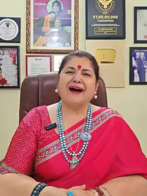 A woman in a red sari with a bindi is sitting in a chair and smiling while the background consists of pictures and an award.