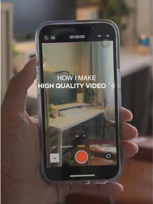 A hand holding a phone that is displaying the phone's camera view of a desk with a white computer screen and other items. The hand is slightly out of focus. Text overlayed on the phone's screen says 'HOW I MAKE HIGH QUALITY VIDEO'.