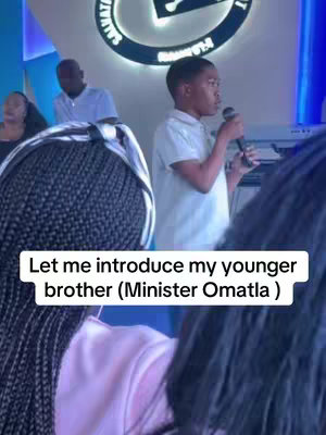 The cover image shows a young Black boy in a white collared shirt speaking into a microphone. He is positioned in front of a blurred background, likely a stage or platform. Above him, a circular logo is partially visible, and text overlayed on the image says, "Let me introduce my younger brother (Minister Omatla )"