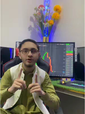 The video shows a young man in a well-lit room, sitting in front of a large monitor displaying graphs. He is wearing glasses, a light-colored vest over a green shirt. A floral arrangement is visible behind him. He is gesturing with his fingers.