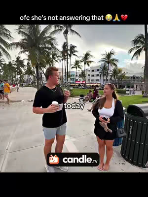 A medium shot of a man and a woman standing on what appears to be a beach walkway. The man on the left is wearing a black t-shirt, light grey shorts, and is holding a white cup. The woman on the right is wearing a white top, a black open jacket, and a black skirt. They both appear to be facing each other. The backdrop is a beach scene with palm trees, the ocean, and a cloudy sky. In the foreground, there is a trash can and the logo of cande.tv.