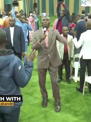 The image shows a crowd of people in a large room. A man wearing a suit is speaking into a microphone in the foreground. People in the background are raising their hands, and the lighting suggests a stage or event setting.