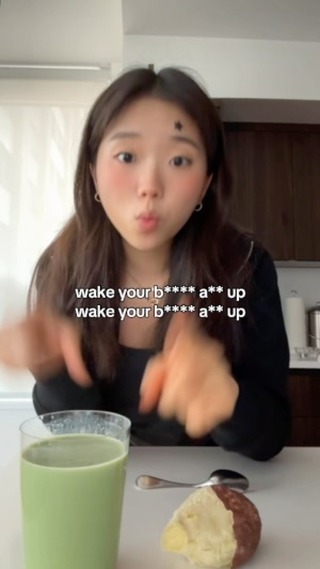 A young woman with long brown hair is shown in a kitchen setting, looking directly at the camera with a slight pout. She has light makeup on, including blush, and a small star-shaped beauty mark above her right eyebrow. The text overlay reads "wake your b**** a** up" in a white, bold font. In the foreground, there is a glass of green liquid, a spoon, and a piece of bread with a dark, round filling.