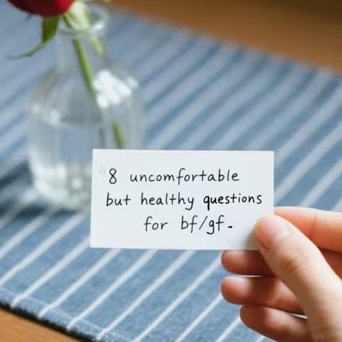 A hand holds a white rectangular card with handwritten text in black, against a backdrop of a blue and white striped tablecloth, with a miniature glass vase holding a red rose slightly out of focus in the background.