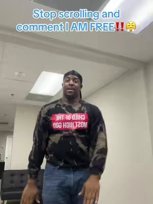 A man is standing in a room, with a caption overlaid on the image. The man is wearing a black shirt with a graphic on it and blue jeans, and a black durag. Above him is text that reads stop scrolling and comment I AM FREE.