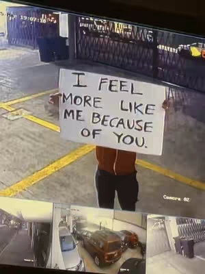 The cover image shows a person holding a white sign with the message 'I FEEL MORE LIKE ME BECAUSE OF YOU.' The setting appears to be an outdoor parking area with a security camera visible at the top.  Below the sign, there are several smaller frames showing different views of the parking area, including parked cars and a gated entrance. The overall composition is captured from a security camera angle.