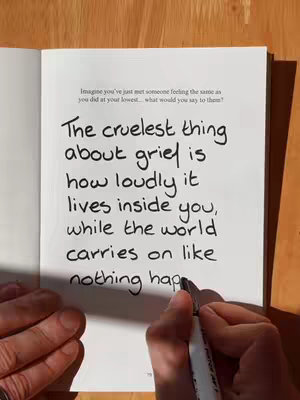 An open book with a handwritten message on one of its pages. A hand with a black marker is writing on the page. The text is in the center, with a wooden background in the top left and right corners.