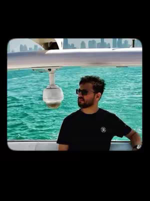 The image shows a man in sunglasses and a black t-shirt standing on a boat. He is facing the side, with a water background and buildings in the distance. A camera is mounted above.