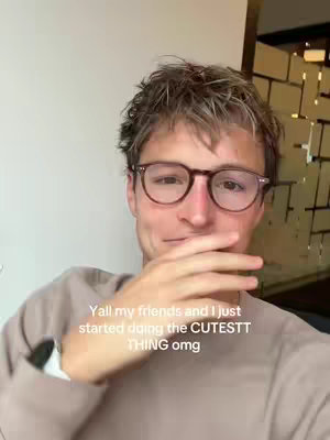 A young man with light brown curly hair and glasses is holding up his hand, which partially obscures his face. He is facing the camera, smiling slightly while sitting in front of a neutral white and golden background.