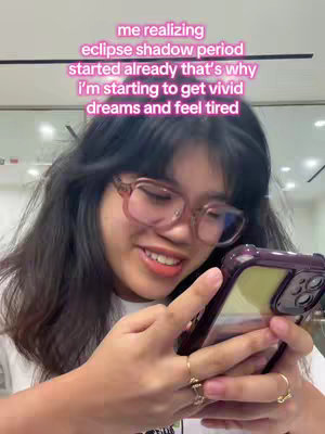 A young woman with dark hair and glasses is holding a smartphone in her hands. She appears to be looking at the screen with a smile, and the background is slightly blurred. Overlayed text in pink, indicates the eclipse season has begun and the feeling of vivid dreams and tiredness.
