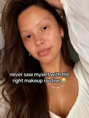 A self-shot video; a young woman with long brown hair is the main focus, her face is centered. She is looking towards the camera. Text is overlaid over her image across the center that reads "never saw myself with the right makeup routine". She is wearing a white top, and a silver necklace with rhinestones sits on her neck. The background is simple, and the lighting appears to be soft and natural.
