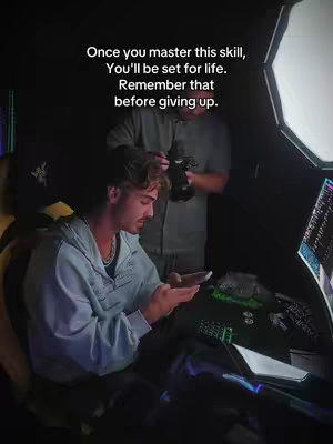 A man is featured in a dimly lit room, likely a home office or studio with screens. He is seated, looking down at a phone in his hands, with a second person partially visible behind him holding a camera or recording device. Text overlayed conveys motivational, inspirational content.