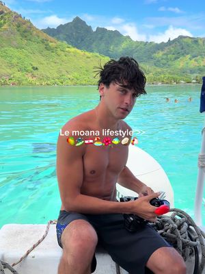 A shirtless man sits on a boat, looking at the camera. The background is a stunning tropical scene with mountains. Text and emojis are overlaid at the top.