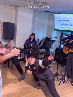 The image shows a group of people in a room, likely a church or meeting hall. The foreground features a person kneeling and extending their arm. Other individuals are visible, some standing and possibly singing. A keyboard is present, and a screen displays text, suggesting a performance or service is in progress. The lighting seems to be indoor, possibly stage lighting.