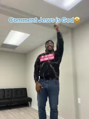A man in a black and white patterned sweatshirt stands in an empty room with one arm raised. Text overlayed on the image says, "Comment Jesus is God " and includes a smiling face emoji. The man is wearing blue jeans and a black cap. The background is a plain wall lit with overhead recessed lighting.