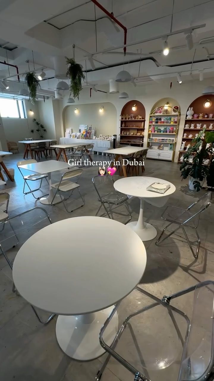 An interior shot of an open-plan space. White tables and chairs are set up, with clear and white chairs scattered throughout. There is a wall with multiple decorative arch-shaped shelves. Hanging plants and pendant lights also seen. The overall ambiance is bright and airy.