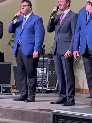 Vertical shot of three men in suits, singing into microphones. The first man on the left wears a blue suit, the middle man wears a dark grey suit, and the right side of the frame shows a hint of the third man's blue suit as well. They are on stage, with basic audio equipment visible.