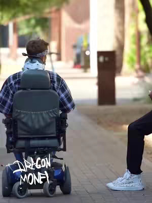 A medium shot shows a person from the back in a wheelchair, with the text 'DRAGON MONEY' overlaid at the bottom. The person is wearing a plaid shirt and a bandana and is facing away from the camera. Another person, wearing black pants and white sneakers, is seen standing to the right. The background is a pathway with trees.