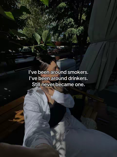 A selfie-style image featuring a person in a casual outdoor setting. The person is in the lower part of the frame, with a top-down perspective. The background shows greenery, possibly a garden or park, with hints of sunlight filtering through. The person is centrally positioned, wearing a white shirt over a black top and the image has overlaid text.
