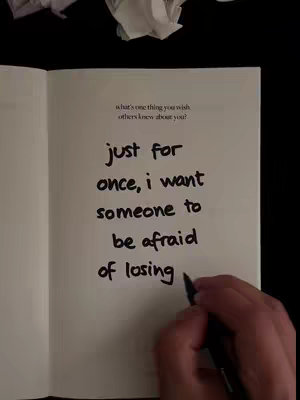 An open book lays face up, with black text hand-written across the page. A hand with a pen is in the bottom right-hand corner, with the pen tip in the process of writing on the page. The book is white, with the black text contrasting against it. A crumpled piece of white paper is in the top left corner, and the background is a simple black.