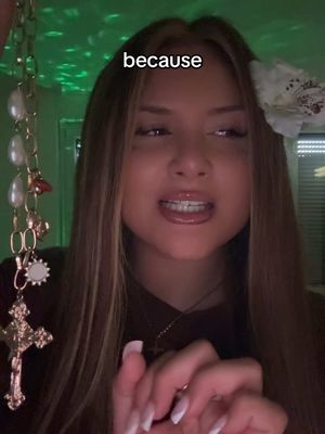 A young woman with long brown hair and subtle makeup is the central focus, positioned centrally in the frame. She is wearing a dark maroon top and a delicate cross necklace. A jeweled chain with pearl and gold pendants hangs on the left of the frame, and a white flower is placed in her hair. The background appears to be a dimly lit room, with spots of green light. The woman is looking up with parted lips, as if mid-sentence.