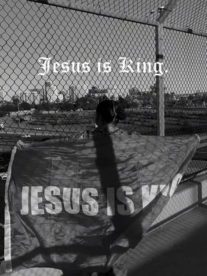 A black and white image shows a person facing away from the camera, holding up a banner that reads “JESUS IS KING.” The background includes a chain-link fence, some buildings, and cityscapes, and the sky is visible.