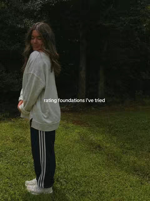 A woman stands in a grassy area with a dark, blurred forest in the background. She's wearing an oversized light-colored sweatshirt, dark blue track pants with white stripes, and white sneakers. She's angled to the side, looking towards the camera.