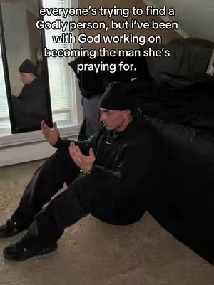 A man in a black hoodie and beanie kneels on the floor with his hands up in a prayer-like gesture. He's in a room with a window and a mirror reflecting his image. Text is layered over the image.