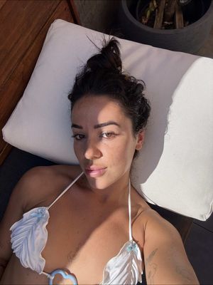 A selfie-style shot of a woman reclining on a white pillow in a sunlit outdoor setting. She is wearing a white bikini top and has dark hair styled in a bun. The lighting suggests a sunny day, and a plant pot is visible in the background.