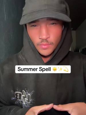A selfie-style shot features a young man wearing a black hoodie and a dark cap, set against a blurred dark background. A text overlay states Summer Spell with sun, star and crescent moon emojis . The man is looking down with his hands clasped in front of him. The lighting is focused on his face, creating a warm, natural glow.