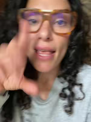 A close-up shot of a woman with curly hair and large glasses, wearing a gray shirt. She is looking directly at the camera with her hand raised, seemingly interacting with the viewer. The image has a casual, personal feel.