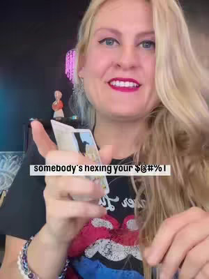 A woman with long blonde hair holds tarot cards, smiling at the camera against a backdrop of a dimly lit space with a small figurine on a shelf. The text overlay reads, "somebody's hexing your $#@%!"