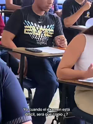 A classroom setting with a dark-haired young man at the front desk, appearing to consider something during an exam. The camera angle shows him from mid-torso up, and the angle is slightly from the side. There are other students visible in the background. The image has a muted color palette, with the man wearing a black t-shirt, blue jeans and sitting in front of a wooden desk.