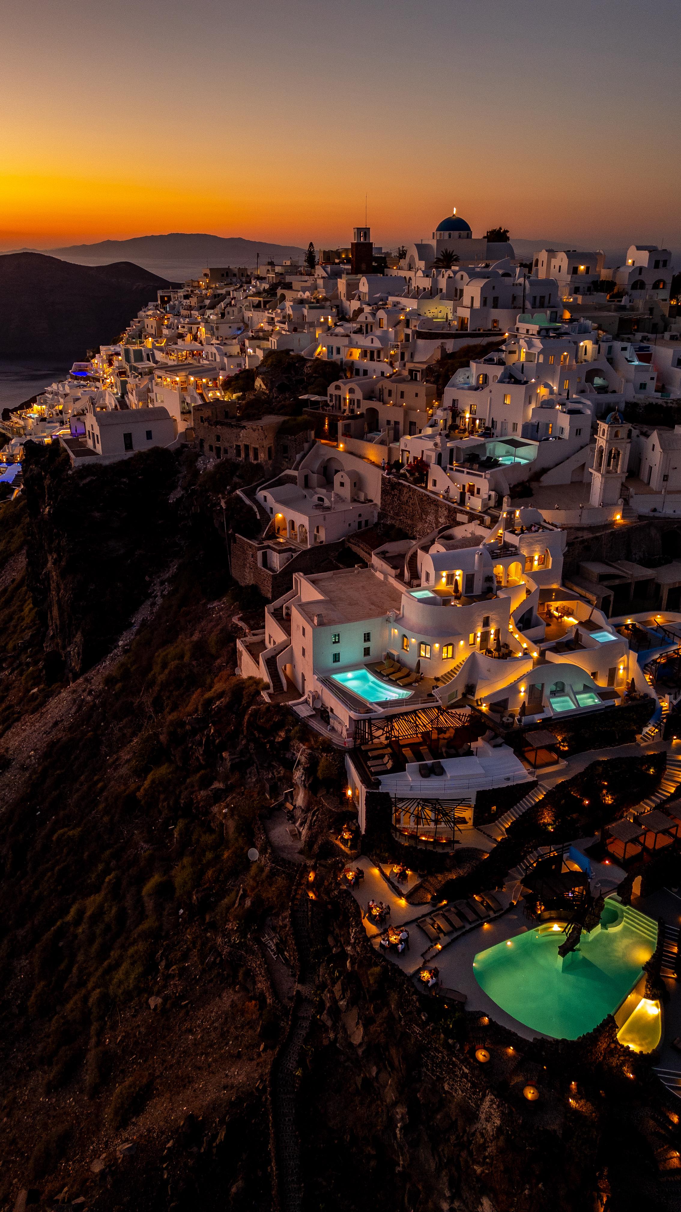 An aerial view captures a picturesque Santorini cityscape at dusk, dominated by white-washed buildings with blue-domed churches. The scene is bathed in the warm hues of the setting sun, with pools illuminated and the sea visible in the distance.