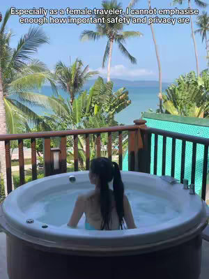 The image shows a woman in a jacuzzi, her back to the camera, set against a lush tropical backdrop. Palm trees and dense green foliage surround a balcony with a wooden railing, offering a view of the ocean and a clear blue sky. The jacuzzi is filled with clear water and bubbles, and the woman is wearing a light-colored swimsuit.
