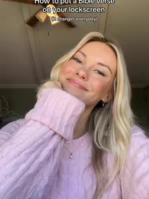 A woman with long blonde hair and a pink sweater is positioned in the image, with a soft smile and hand touching her face. Above, text in a white box states, 'How to put a Bible verse on your lockscreen, changes everyday'. The background is slightly blurred, with a ceiling fan visible.