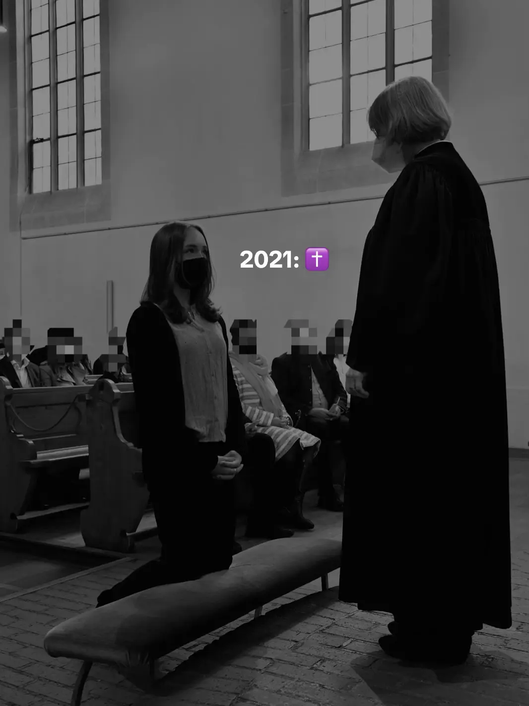 A black and white image shows a woman kneeling before another person in a chapel. The woman kneeling is wearing a black face mask. The image is framed with a lot of empty space that is filled with blurred figures, indicating the presence of a congregation.