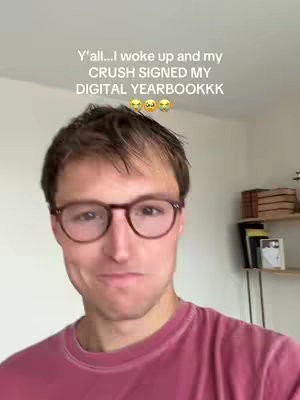 A man with short brown hair wearing glasses is looking at the camera with a slight smile. He is wearing a red t-shirt, and the background is a plain white wall with a shelf of books on the right-hand side.