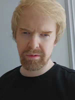 A man with light blonde hair and a matching beard is shown close-up. He wears a black t-shirt and has a somewhat stern or questioning expression. The background is plain and light-colored, possibly a wall or door.