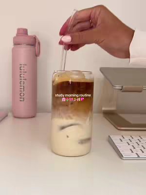 The image shows a well-lit, aesthetic desk setup featuring a glass of layered iced coffee with a straw, a pink Lululemon water bottle, a laptop stand, a laptop, and a keyboard. A hand with pink nail polish holds the straw above the drink. The text "study morning routine" is overlaid on the glass, along with various emojis.