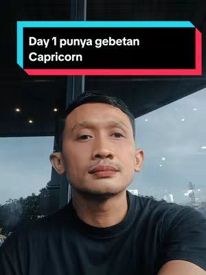 The cover image features a man in a black t-shirt, positioned in the lower half of the frame. Above him, there's a rectangular box with the text "Day 1 punya gebetan Capricorn" in white against a black background, framed by a red border at the top and a blue border at the bottom. The background behind the man appears to be a window with a cityscape visible through it.