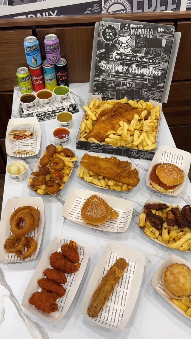 A top-down view of various dishes from a fish-and-chips shop arranged on a white surface. A large box labeled “Mother Hubbard’s Super Jumbo” sits in the upper right, partially obscuring the background. Several servings of fish and chips, along with other items like burgers, onion rings, and sauces, fill the frame, giving the impression of a full menu display. Cans of soda and small containers of dipping sauces are placed around the food.