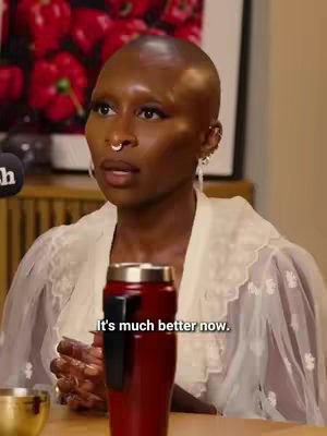 A woman with a shaved head and a nose ring is seated and speaking. She is wearing a white top with a frilly collar. A travel mug with a black handle and a wooden table are directly in front of her. The background shows a framed piece of art and a wooden wall.