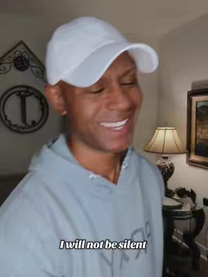 A middle-aged Black man wearing a white baseball cap and a gray hoodie smiles widely, revealing white teeth. In the background, a room with a decorative wall element, a framed picture, and a lamp is visible. Text overlayed at the bottom reads, "I will not be silent."